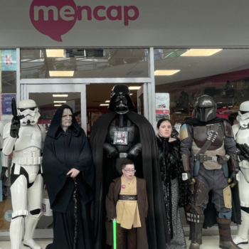 A group of Star Wars characters, including Darth Vader, Emperor Palpatine, a young Jedi, two Stormtroopers, a Mandalorian, and other cosplayers, stand in front of a Mencap charity shop. They are dressed in detailed costumes and pose together outside the storefront for a charity event at Walkden Shopping Centre.